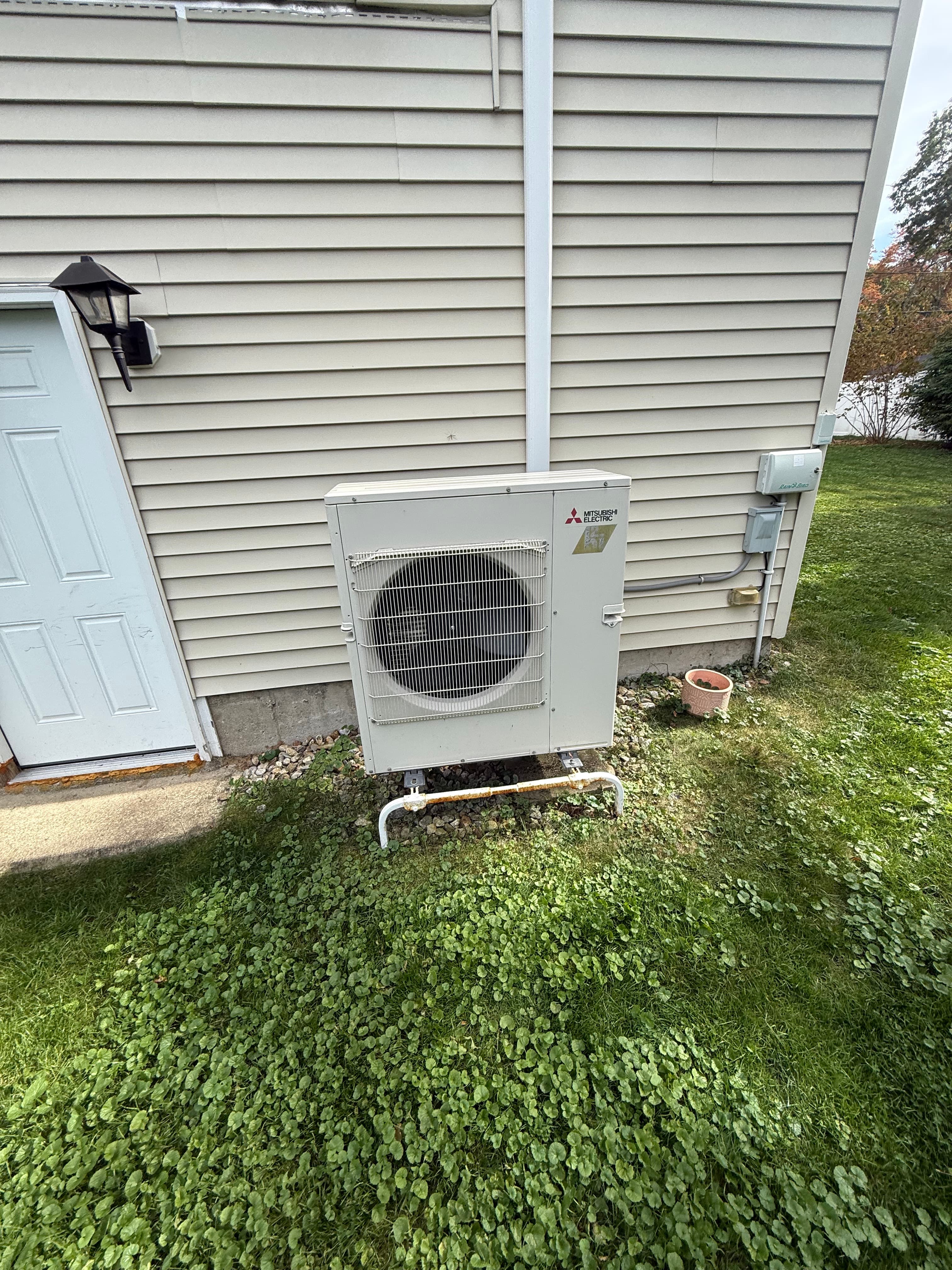 Mitsubishi Electric outdoor unit mounted on a beige-sided house wall next to green grass.